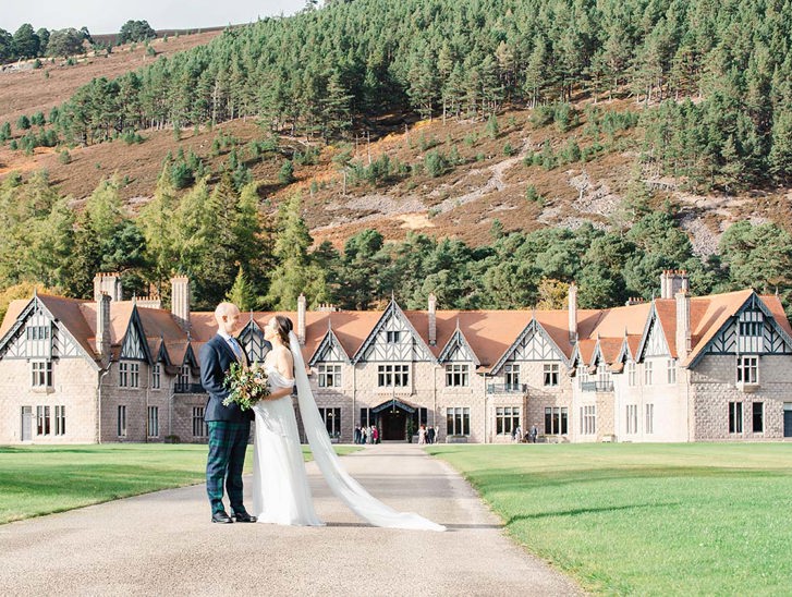 bride and groom standing in front of large country house with Scottish hills surrounding them